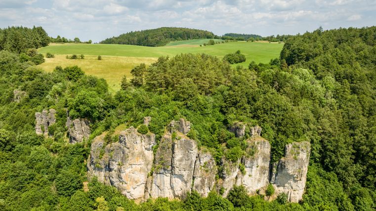 An impressive rock formation surrounded by lush greenery and gentle hills. The sky is partially cloudy, giving the landscape a peaceful atmosphere.