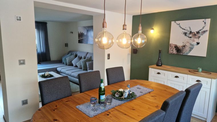 Ein elegant eingerichteter Essbereich mit einem runden Holztisch und modernen Hängelampen. Im Hintergrund ist ein gemütliches Wohnzimmer mit einer grauen Couch sichtbar.