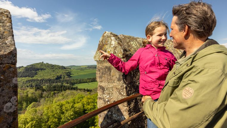 Ein Mann und ein Kind auf der Kasselburg, genießen den Ausblick auf die grüne Eifellandschaft unter blauem Himmel.
