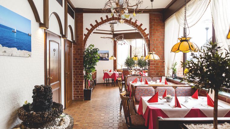 In einem Raum mit mediterranem Stil stehen mehrere Tische mit Stühlen und Sitzbänken in rot, der Boden ist befließt und im Raum stehen mehrere Pflanzen