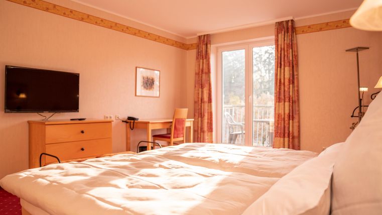 A bright room with a large bed and a television. Large windows let in a lot of light and offer a view of the balcony.