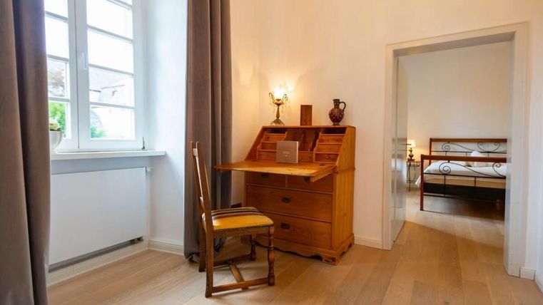 Ein helles Zimmer mit einem Schreibtisch und einer Holzstuhl. Im Hintergrund ist ein weiterer Raum mit einem Bett sichtbar.