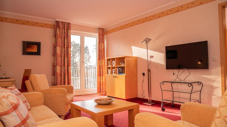 A bright living room with comfortable sofas and a coffee table. A television hangs on the wall, and large window doors provide plenty of natural light.