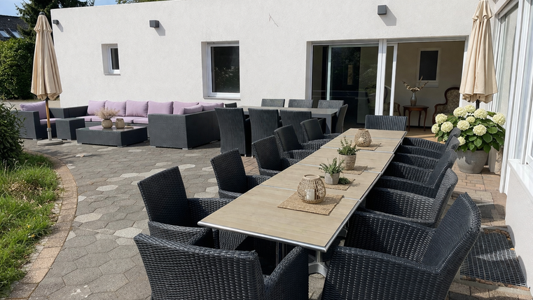 Une espace extérieur élégant avec une longue table et des chaises confortables. En arrière-plan, il y a des sièges confortables et un joli jardin.