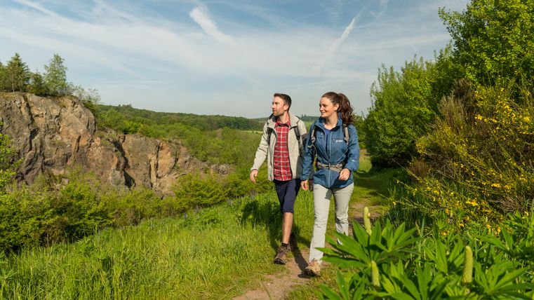 Zwei Wanderer inmitten der Natur auf einem Wanderweg. Im Hintergrund ist eine große Felswand zu sehen. Hierbei handelt es sich um den Vulkan Steffelnkopf.