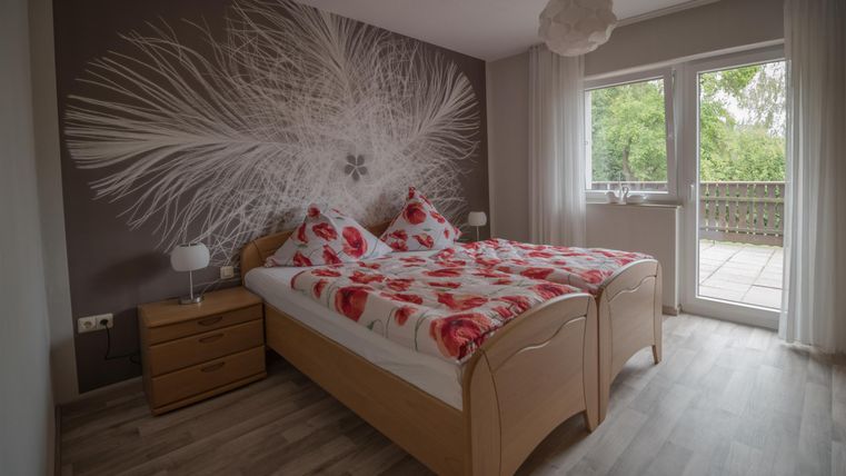 A modern bedroom with a large bed and colorful cushions. Large windows ensure plenty of natural light and provide access to a balcony.