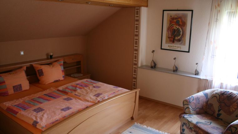 Ein gemütliches Schlafzimmer mit einem Doppelbett, bunten Kissen und einem Sessel. Das Zimmer hat Holzdetails und große Fenster, die viel Licht hereinschicken.