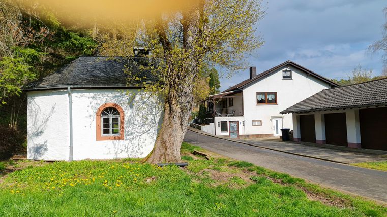 A picturesque house with a large tree next to it. In the background, additional buildings and a quiet path can be seen.