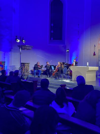 Musiker an Streichinstrumenten musizieren vor dem Kirchenaltar. Davor die Sitzbänke voller Besucher.