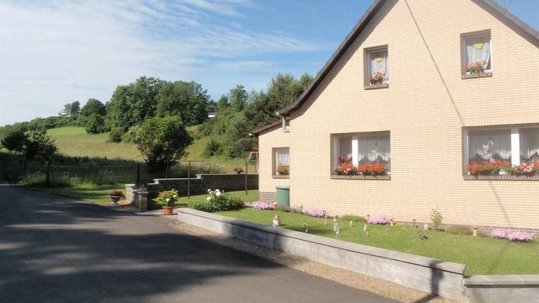 Ein charmantes Haus mit einem hellen Außenbereich und bunten Blumen. Im Hintergrund sind grüne Wiesen und Bäume zu sehen.