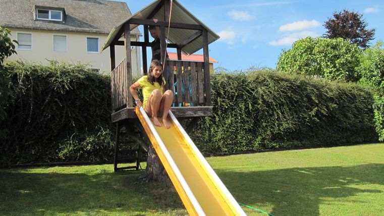 Ein Kind rutscht von einer gelben Rutsche in einem Garten. Im Hintergrund ist ein Spielhaus und grüne Bäume zu sehen.