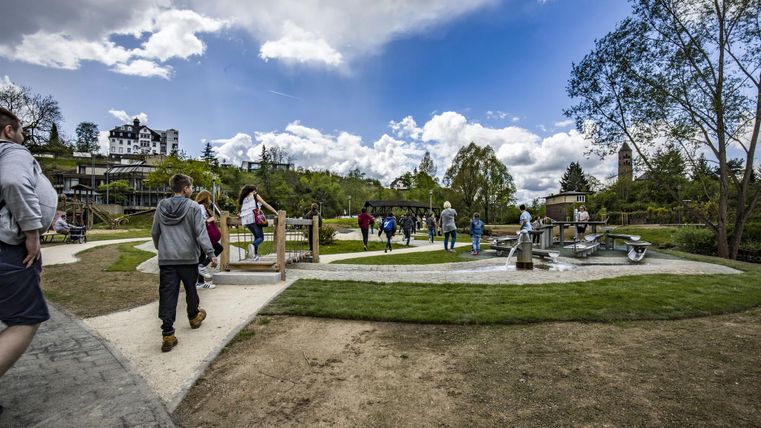 Menschen spazieren auf einem Spielplatz im Gerolsteiner Kurpark. Im Hintergrund sind Bäume, ein Gebäude und ein Turm zu sehen. Der Himmel ist bewölkt.