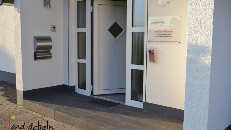 Ein einladender Eingangsbereich mit einer modernen Haustür und einer klaren Fassade. Neben der Tür befindet sich ein Briefkasten und eine Informationsschilder.