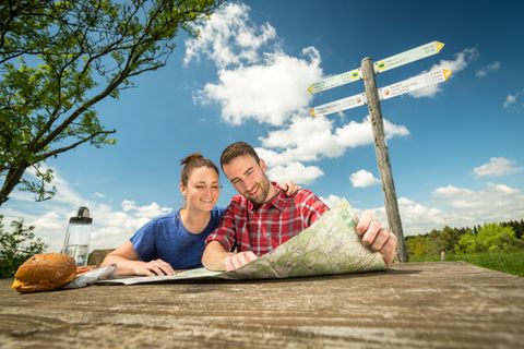 Ein Mann und eine Frau sitzen auf einer Rastbank inmitten der Natur und studieren eine Wanderkarte auf dem Tisch. Daneben liegt Proviant auf dem Tisch.