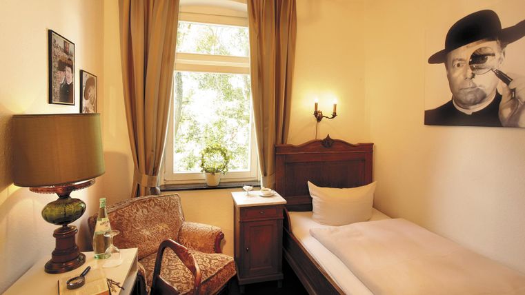 Une chambre confortable avec un lit simple et une chaise. Un portrait d'un homme est accroché au mur, et la fenêtre laisse entrer beaucoup de lumière naturelle.