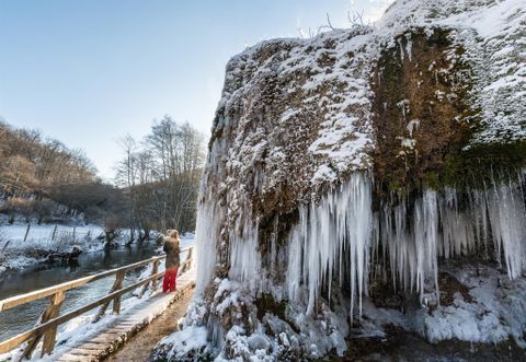 Gefrorener Nohner Wasserfall mit Eiszapfen, eine Person auf einem Steg, winterliche Landschaft mit Schnee und kahlen Bäumen