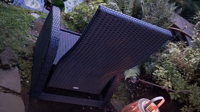 Een comfortabele stoel staat naast een vijver, omringd door planten. Een lantaarn zorgt voor zacht licht in de schemering.