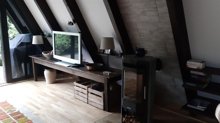 Ein modernes Wohnzimmer mit schrägen Wänden und großen Fenstern. Es gibt einen Fernseher, einen Holzofen und gemütliche Möbel.