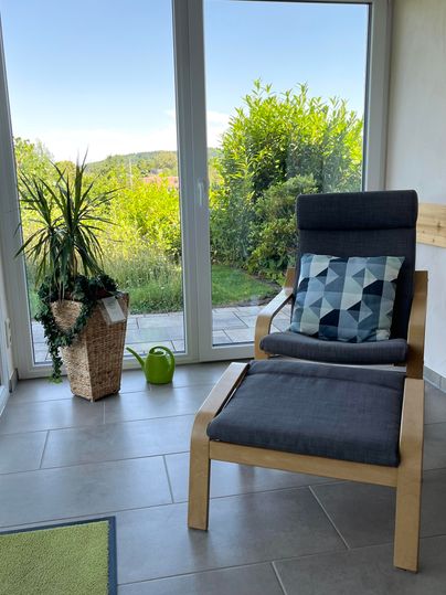 Un fauteuil confortable se trouve devant de grandes fenêtres donnant sur un jardin verdoyant. À côté du fauteuil se trouve un pot avec une plante et un petit arrosoir.