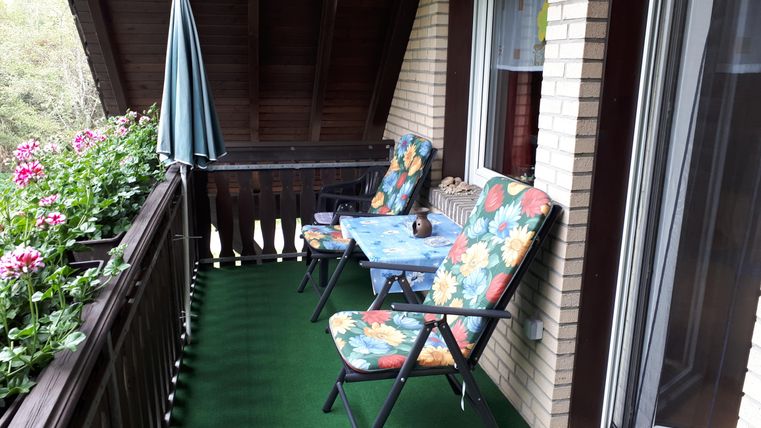 Ein gemütlicher Balkon mit bunten Stühlen und einem Tisch. Über die Geländer sind Blumen angebracht, und ein Sonnenschirm steht bereit.