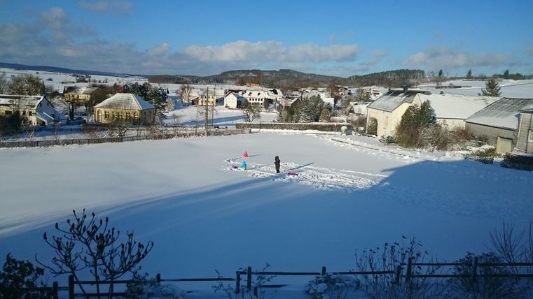 Eine verschneite Landschaft mit einem großen weißen Feld und einem einzelnen Menschen, der darauf arbeitet. Im Hintergrund sind Häuser und Bäume unter einem klaren blauen Himmel zu sehen.