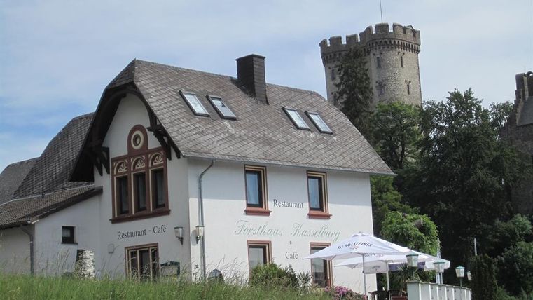 Weißes Gebäude des Forsthauses Kasselburg mit Café und Restaurant, im Hintergrund ein steinerner Turm mit deutscher Flagge.