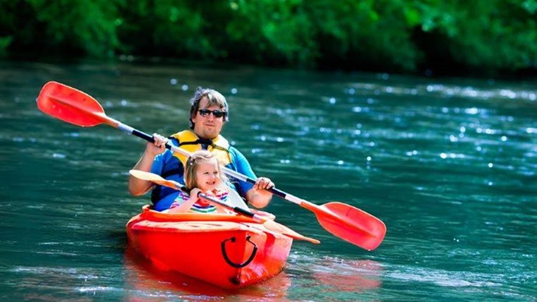 Ein Vater und seine Tochter paddeln fröhlich in einem roten Kajak auf einem ruhigen Fluss. Umgeben von grünen Bäumen genießt die Familie die Natur.