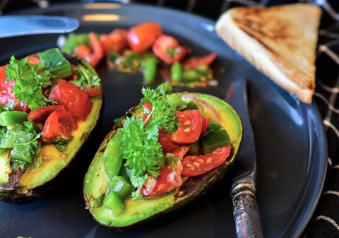 Gefüllte Avocadohälften mit Tomaten und Paprika auf einem Teller, daneben ein Stück Toast.