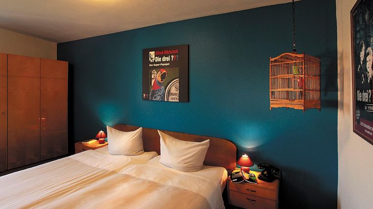 Une chambre confortable avec deux lits simples et un mur bleu. Il y a une commode, des lampes de chevet et une cage décorative au mur.