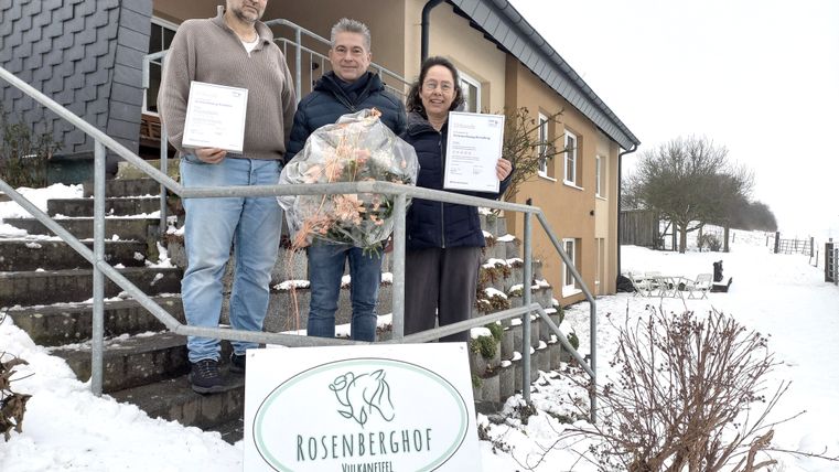 Das Bild zeigt drei Personen, die vor einem Haus im Schnee stehen. Die beiden äußeren Personen halten Urkunden in der Hand, während die mittlere Person einen Blumenstrauß trägt. Vor ihnen steht ein Schild mit der Aufschrift "Rosenberghof Vulkaneifel Ferienwohnung" und einem QR-Code. 