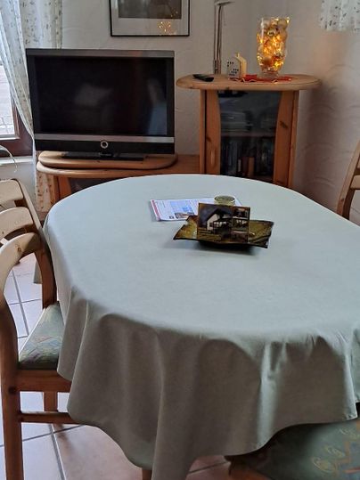 Ein Esszimmer mit einem runden Tisch, der mit einer hellen Tischdecke gedeckt ist. Im Hintergrund stehen ein Fernseher und ein Kamin, umgeben von einer gemütlichen Atmosphäre.