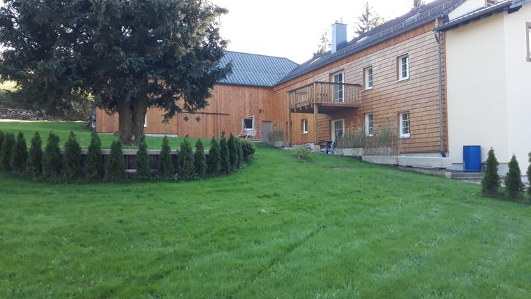 Ein gepflegter Garten mit grünem Rasen und kleinen Bäumen. Im Hintergrund stehen zwei Häuser mit Holz- und Putzfassade.