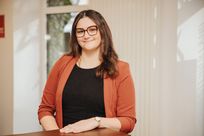 Eine Frau steht hinter einem Tresen in einem hellen Büroraum und lächelt in die Kamera. Hinter ihr einige Fenster.