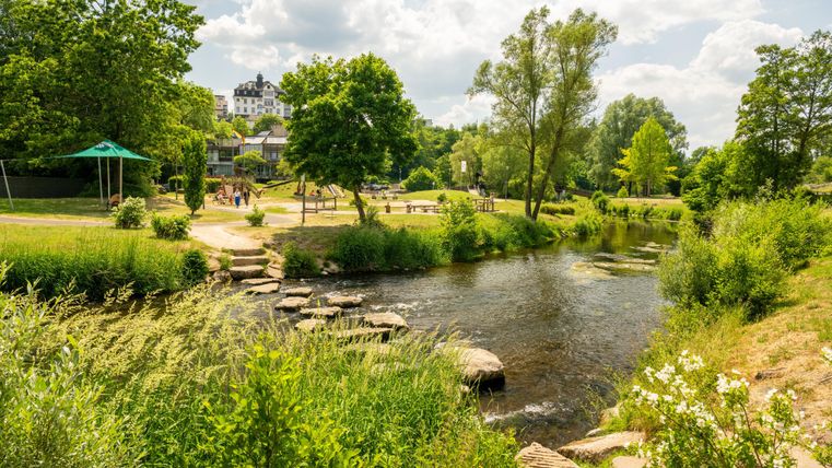 Ein idyllischer Park mit Fluss, Bäumen und einem Spielplatz. Menschen spazieren und genießen die Natur. Im Hintergrund ein großes Gebäude.