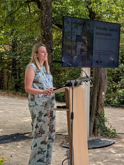 Eine Frau steht an einem Podest und hält eine Rede. Neben ihr zeigt ein Bildschirm eine Präsentation zum Thema "Virtuelle Tourist-Information"