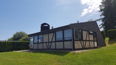 Modernes Ferienhaus mit großen Fenstern und Fachwerkdetails, umgeben von grüner Wiese und Bäumen, unter blauem Himmel.