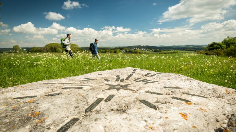 Zwei Wanderer auf einem Feldweg mit einem markierten Stein im Vordergrund.