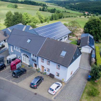Een modern gebouw met zonnepanelen op het dak, omringd door de natuur. Op de voorgrond zijn meerdere auto's en een betegelde parkeerplaats te zien.