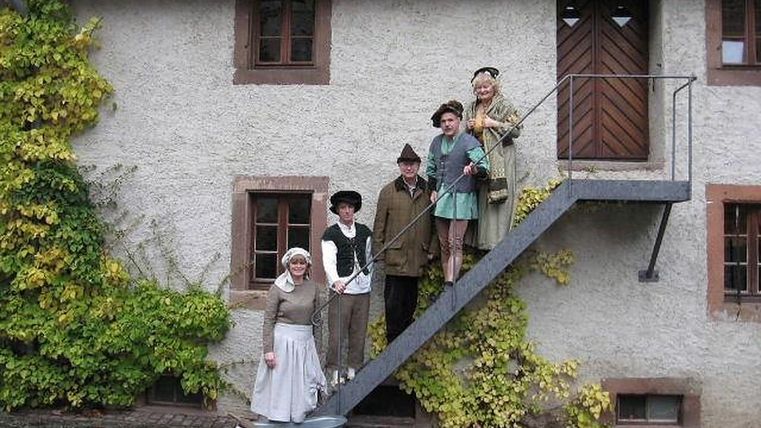 Personen in historischen Kostümen stehen auf einer Treppe vor einem alten Gebäude mit Efeu bewachsen.