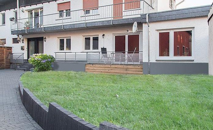 Een modern huis met een verzorgde tuin en een terras. De gevel is licht en heeft enkele ramen met rode jaloezieën.