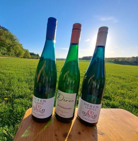 Drei Flaschen Wein stehen auf einem Holztisch, umgeben von einer grünen Wiese. Die Sonne scheint klar und beleuchtet die Szene.