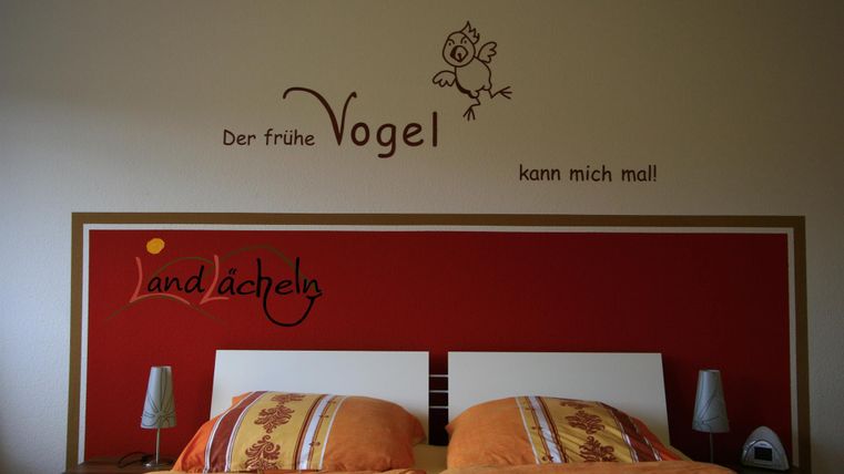 Ein gemütliches Schlafzimmer mit zwei Betten und bunten Kissen. An der Wand steht der Spruch „Der frühe Vogel kann mich mal“.