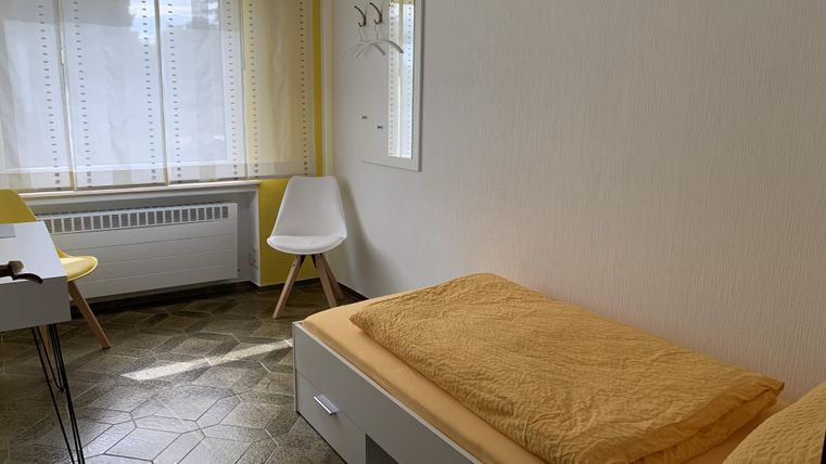Ein einfaches Zimmer mit einem einzelnen Bett und einer gelben Bettdecke. Es gibt einen Tisch, einen Stuhl und viel Tageslicht durch das Fenster.