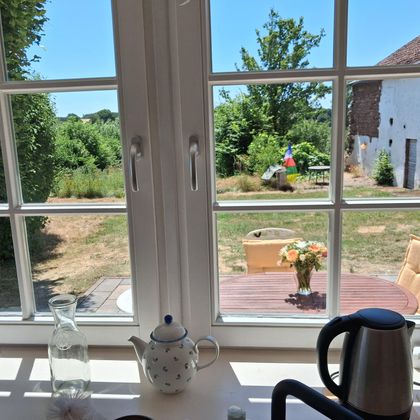 Ein Blick aus einem Fenster auf einen Garten mit sonnigem Wetter. Im Vordergrund sind einige Kaffeetassen und eine Kanne zu sehen.