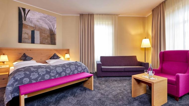 Ein modernes Schlafzimmer mit einem Doppelbett, einem gemütlichen Sofa und einem pinken Sessel. Die Fenster lassen viel Licht herein und die Einrichtung ist geschmackvoll und einladend.