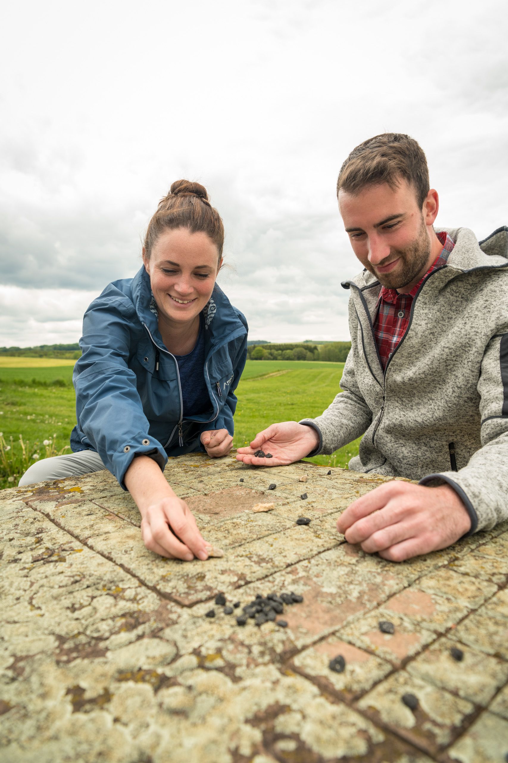 Ein Mann und eine Frau sitzen draußen an einem Steintisch und spielen ein Brettspiel mit kleinen Steinen.