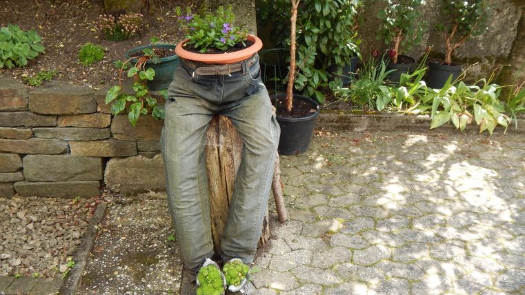 Eine kreative Gartendekoration zeigt eine Person aus alten Hosen und Stiefeln, mit einem Blumentopf auf dem Kopf. Umgeben von Pflanzen fügt sie einen charmanten Akzent zum Garten hinzu.