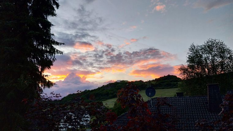 Een schilderachtige zonsondergang boven zachte heuvels. De wolken zijn in warme kleuren gekleurd en omringd door bomen en een woning.