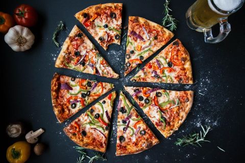 Eine frisch gebackene Pizza, in Stücke geschnitten, liegt auf einem dunklen Tisch. Neben der Pizza steht ein Glas Bier und einige frische Zutaten.