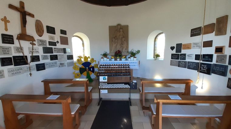 Innenraum der Kapelle Wahlhausen mit Holzbänken, Kerzenständer und Danktafeln an den Wänden. Ein Kruzifix und religiöse Symbole sind sichtbar.